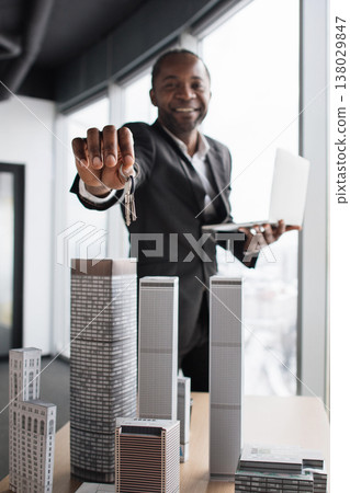 A smiling African man in a suit holds keys and a laptop, standing behind miniature skyscrapers, symbolizing real estate A smiling African man in a suit holds keys and a laptop, standing behind miniature skyscrapers, symbolizing real estate 138029847