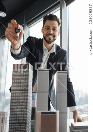 A smiling agent presents keys over a scale model of high-rise buildings in a modern office setting with city views and daylight windows indoors A smiling agent presents keys over a scale model of high-rise buildings in a modern office setting with city views and daylight windows indoors 138029857