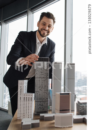A smiling businessman in a suit stands by a table displaying various architectural models of buildings, with a large window behind him A smiling businessman in a suit stands by a table displaying various architectural models of buildings, with a large window behind him 138029879