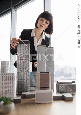 A woman in a suit examines architectural models of buildings, showcasing urban planning and design concepts A woman in a suit examines architectural models of buildings, showcasing urban planning and design concepts 138029892