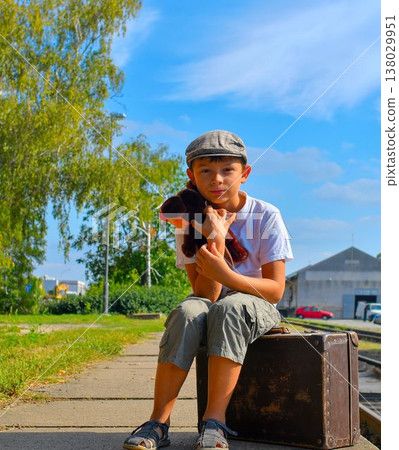 A cute little boy on a train station platform. A happy little boy holds a plush toy and sits on an old suitcase. The concept of travel, vacation, and a carefree childhood. Retro and vintage vibe. A cute little boy on a train station platform. A happy little boy holds a plush toy and sits on an old suitcase. The concept of travel, vacation, and a carefree childhood. Retro and vintage vibe. 138029951
