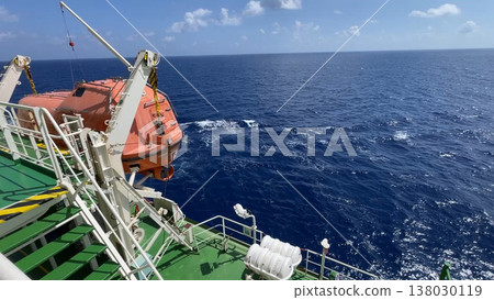 A bright orange fast rescue boat secured on its launching davit on the deck of a commercial vessel, with a background of choppy ocean waves during transit in windy maritime conditions 138030119