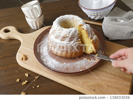 Homemade Gugelhupf bundt cake with powdered sugar on wooden cutting board. Hand cutting slice of fresh pastry with knife in rustic kitchen setting. Homemade Gugelhupf bundt cake with powdered sugar on wooden cutting board. Hand cutting slice of fresh pastry with knife in rustic kitchen setting. 138033036