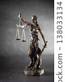 A bronze statue of Justice stands against a dark, textured background with dramatic side lighting. The figure holds scales symbolizing justice in one hand and a sword in the other, her eyes covered by 138033134