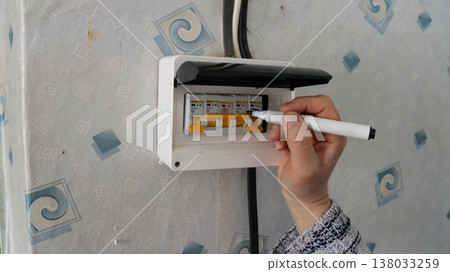 A hand holding a black marker applies labels to the circuit breakers in a residential distribution board, and also marks the meter mounted on the wall. The breakers for various electrical circuits 138033259