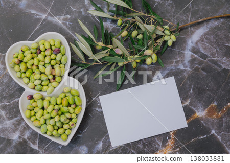 Healthy green olives in heart shaped white plate with tree branch, Mediterranean superfood rich in vitamins and minerals, blank paper for copy space 138033881