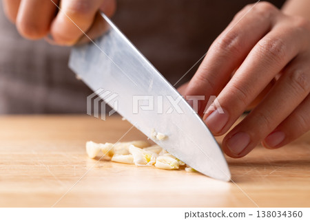 Hand holding a knife and mincing fresh garlic on a wooden cutting board, prepared for cooking Hand holding a knife and mincing fresh garlic on a wooden cutting board, prepared for cooking 138034360