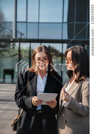 Two women engage in discussion while using a tablet in a modern office setting during a sunny day in the city 138034505