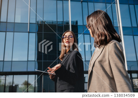 Confident businesswoman discusses ideas with a model in front of a modern office building during the day in a city setting 138034509