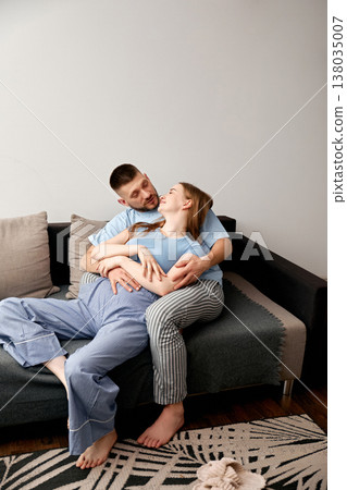 Couple sits together on a couch in a cozy living room while enjoying their time and sharing smiles in the morning 138035007