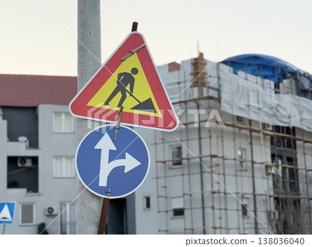 Roadwork warning sign and direction arrow mounted on pole near apartment construction site. Traffic regulation, street safety, detour guidance, urban development, temporary navigation in changing Roadwork warning sign and direction arrow mounted on pole near apartment construction site. Traffic regulation, street safety, detour guidance, urban development, temporary navigation in changing 138036040
