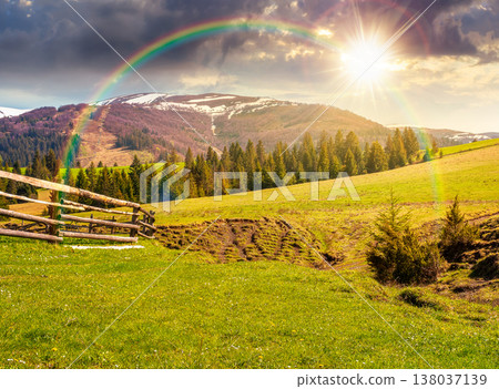 sunset over mountainous countryside landscape in spring. wooden fence and grassy meadow on a green hill. distant mountain ridge with snow capped tops. cloudy sky and rainbow 138037139