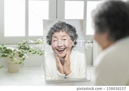 一位80歲的老婦人對著鏡子微笑，輕輕撫摸自己的臉頰。護膚與美容圖片 138038237
