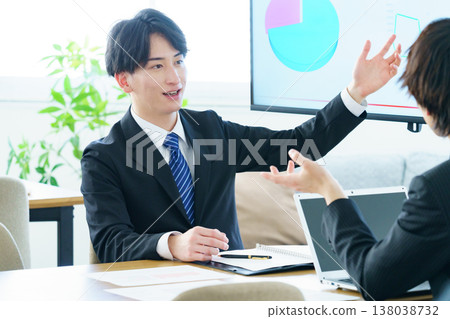 A young man gives a presentation in an office, explaining a graph displayed on a screen. A young man gives a presentation in an office, explaining a graph displayed on a screen. 138038732