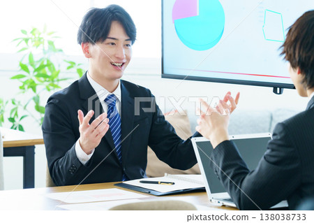 A young man gives a presentation in an office, explaining a graph displayed on a screen. A young man gives a presentation in an office, explaining a graph displayed on a screen. 138038733