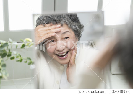 一位80歲的老婦人面帶微笑,雙手輕撫額頭和臉頰,對著鏡子欣賞自己的外表。護膚與美容圖片 一位80歲的老婦人面帶微笑,雙手輕撫額頭和臉頰,對著鏡子欣賞自己的外表。護膚與美容圖片 138038954