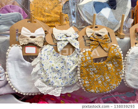 Floral baby bibs and matching bows arranged at a handmade accessories market stand. Infant feeding essentials, nursery styling, gift selection, and boutique textile craftsmanship in soft pastel tones Floral baby bibs and matching bows arranged at a handmade accessories market stand. Infant feeding essentials, nursery styling, gift selection, and boutique textile craftsmanship in soft pastel tones 138039293