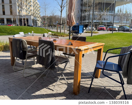 Large communal table with chairs and blankets on a landscaped cafe terrace in sunshine. Outdoor hospitality, social gathering, coworking pause, and modern urban leisure near residential architecture Large communal table with chairs and blankets on a landscaped cafe terrace in sunshine. Outdoor hospitality, social gathering, coworking pause, and modern urban leisure near residential architecture 138039316