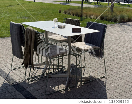 Outdoor cafe table with cushions, blankets, and reserve sign on a sunny terrace. Patio seating, seasonal hospitality, al fresco dining, and comfortable urban leisure beside landscaped lawn Outdoor cafe table with cushions, blankets, and reserve sign on a sunny terrace. Patio seating, seasonal hospitality, al fresco dining, and comfortable urban leisure beside landscaped lawn 138039323
