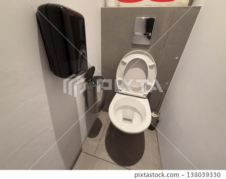 Toilet with open seat, flush plate, bin, and brush in a narrow restroom stall. Sanitation upkeep, bathroom utility, hygiene routine, and compact fixture layout for everyday maintenance Toilet with open seat, flush plate, bin, and brush in a narrow restroom stall. Sanitation upkeep, bathroom utility, hygiene routine, and compact fixture layout for everyday maintenance 138039330