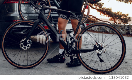Biker prepares for a ride by a dark car at sunset in a parking area by trees Biker prepares for a ride by a dark car at sunset in a parking area by trees 138041082