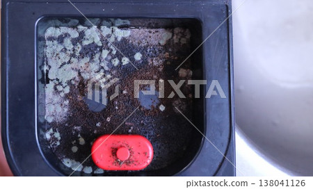 A drip tray from a coffee machine rests on an empty metal sink; inside the tray which belongs to a home coffee machine lie remnants of coffee grounds, covered in fungus and mold 138041126