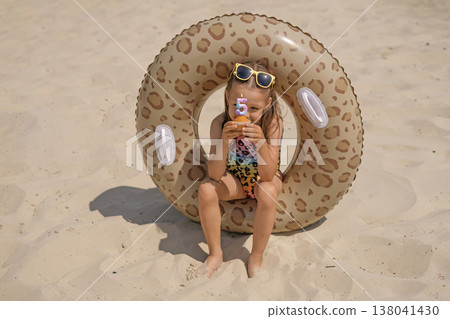 A girl in a swimsuit is sitting on the beach in an inflatable circle holding a cupcake with candles in the shape of the number five, thinking about a wish A girl in a swimsuit is sitting on the beach in an inflatable circle holding a cupcake with candles in the shape of the number five, thinking about a wish 138041430