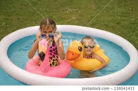 Little girls swim in a mini inflatable pool in the yard on the lawn at home. Children's lifebuoy shaped like flamingos and yellow ducks Little girls swim in a mini inflatable pool in the yard on the lawn at home. Children's lifebuoy shaped like flamingos and yellow ducks 138041431