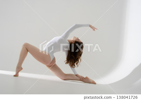 Flexible woman in a white leotard performing a graceful, dynamic stretch on a clean white background. Flexible woman in a white leotard performing a graceful, dynamic stretch on a clean white background. 138041726