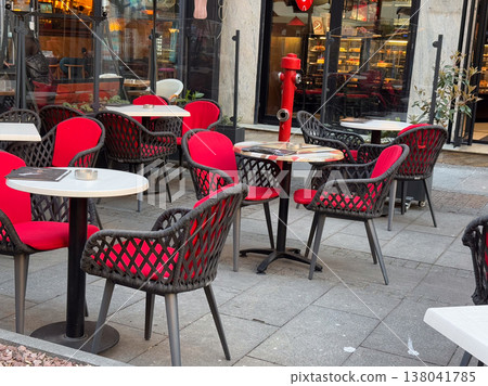 Cafe terrace with red chairs and black woven frames near storefront and fire hydrant. Urban dining scene, street infrastructure integration, hospitality layout, public space usage, city lifestyle Cafe terrace with red chairs and black woven frames near storefront and fire hydrant. Urban dining scene, street infrastructure integration, hospitality layout, public space usage, city lifestyle 138041785