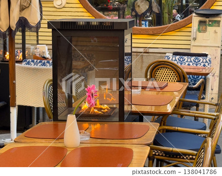 Outdoor cafe heater fireplace with visible flame near table and Gerbera jamesonii flower decoration. Seasonal heating solution, comfort provision, hospitality adaptation, autumn winter dining Outdoor cafe heater fireplace with visible flame near table and Gerbera jamesonii flower decoration. Seasonal heating solution, comfort provision, hospitality adaptation, autumn winter dining 138041786
