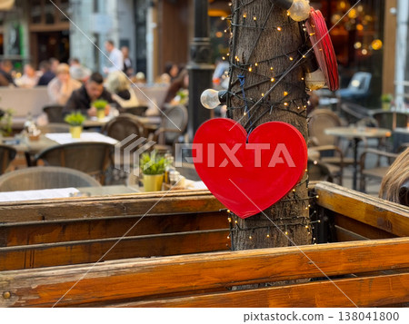 Red heart ornament attached to tree trunk with fairy lights above cafe terrace partition. Romance symbolism, seasonal decoration, date ambiance, hospitality styling, festive street dining atmosphere Red heart ornament attached to tree trunk with fairy lights above cafe terrace partition. Romance symbolism, seasonal decoration, date ambiance, hospitality styling, festive street dining atmosphere 138041800