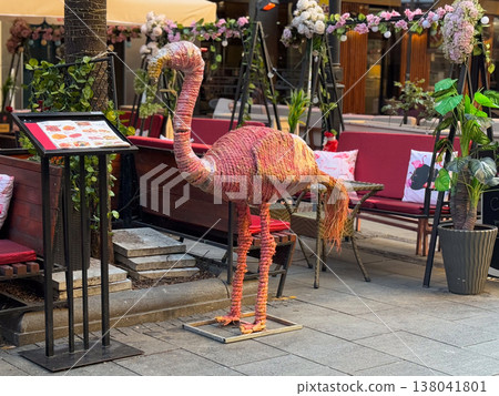 Pink flamingo sculpture beside decorated restaurant terrace and menu stand on city sidewalk. Themed hospitality branding, playful storefront attraction, street marketing, visual identity, customer 138041801