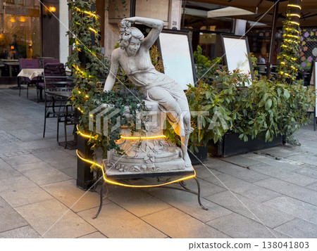 Classical stone sculpture of reclining woman decorated with ivy and light garland near cafe entrance. Artistic decoration, urban aesthetics, cultural reference, hospitality branding, exterior styling 138041803