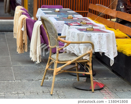 Outdoor cafe seating with wicker chairs and wooden tables along sidewalk partition. Urban hospitality setting, street dining arrangement, small business service, leisure environment, public space Outdoor cafe seating with wicker chairs and wooden tables along sidewalk partition. Urban hospitality setting, street dining arrangement, small business service, leisure environment, public space 138041808