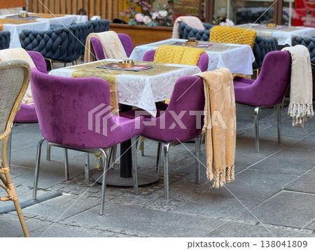 Outdoor restaurant tables with purple chairs and blankets arranged on city terrace. Seasonal seating comfort, textile layering, hospitality service design, customer experience, urban dining 138041809