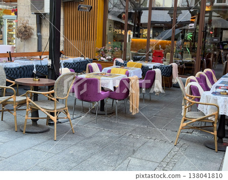 Outdoor restaurant tables with purple chairs and blankets arranged on city terrace. Seasonal seating comfort, textile layering, hospitality service design, customer experience, urban dining 138041810