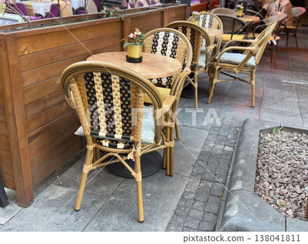 Outdoor cafe seating with wicker chairs and wooden tables along sidewalk partition. Urban hospitality setting, street dining arrangement, small business service, leisure environment, public space 138041811