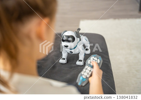 Child controlling robotic dog with remote controller while sitting on sofa at home. Artificial intelligence technology in everyday childhood and smart toy interaction. Child controlling robotic dog with remote controller while sitting on sofa at home. Artificial intelligence technology in everyday childhood and smart toy interaction. 138041832