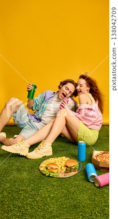 Young couple relaxing on grass with burgers pizza and drinks in bright studio setting. Young couple relaxing on grass with burgers pizza and drinks in bright studio setting. 138042709