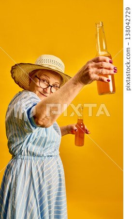 Elderly woman raising bottled drink toward camera making toast on yellow background. Elderly woman raising bottled drink toward camera making toast on yellow background. 138042729