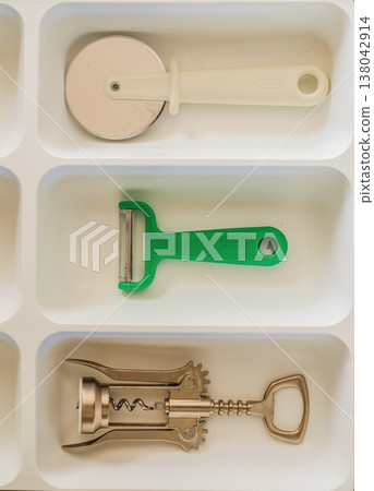 Essential kitchen gadgets: pizza cutter, peeler, and corkscrew neatly organized in a white drawer tray. Essential kitchen gadgets: pizza cutter, peeler, and corkscrew neatly organized in a white drawer tray. 138042914