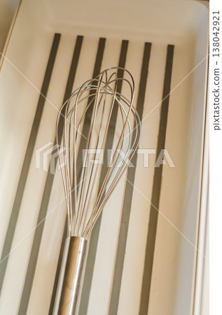A stainless steel kitchen whisk lies in a modern, minimalist white drawer with a striped bottom. Close-up of a cooking utensil. A stainless steel kitchen whisk lies in a modern, minimalist white drawer with a striped bottom. Close-up of a cooking utensil. 138042921