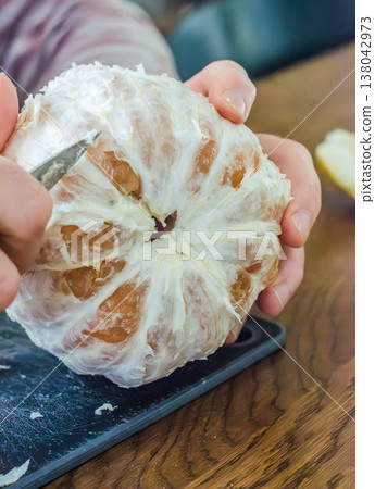 Close-up of hands peeling a large pomelo citrus fruit with a knife over a cutting board. Healthy eating and food preparation. Close-up of hands peeling a large pomelo citrus fruit with a knife over a cutting board. Healthy eating and food preparation. 138042973