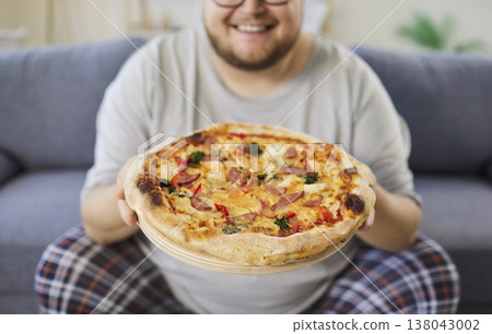 Happy man at home presenting pizza on sofa, casual pajamas, comfort moment Happy man at home presenting pizza on sofa, casual pajamas, comfort moment 138043002