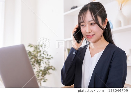 一位二十多歲的女性一邊看著電腦螢幕，一邊用智慧型手機通話。 138044799