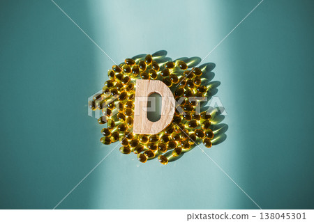 Small wooden letter D stands among yellow translucent supplement capsules on blue surface. Vitamin D deficiency, immune system support, preventative medicine. Small wooden letter D stands among yellow translucent supplement capsules on blue surface. Vitamin D deficiency, immune system support, preventative medicine. 138045301