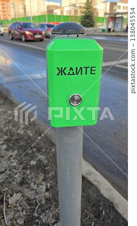 Green pedestrian signal button for crossing the street features Cyrillic text. The metallic button is mounted on a gray post. Green pedestrian signal button for crossing the street features Cyrillic text. The metallic button is mounted on a gray post. 138045554