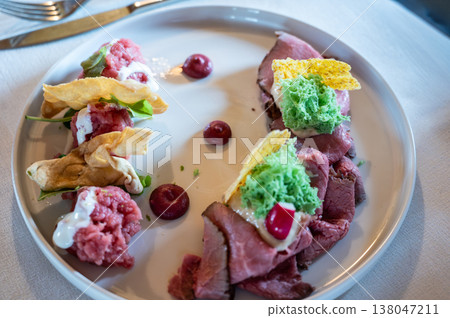 Elegant plated appetizer featuring raw beef tartare and thin slices of roast beef, complemented by green sponge, crispy elements, creamy sauce, and delicate berry drops on a white plate 138047211