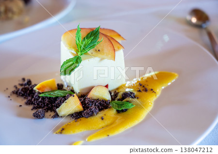 White panna cotta dessert elegantly plated with fresh peach slices, mint leaves, chocolate crumble, and a vibrant mango sauce, representing gourmet culinary art 138047212